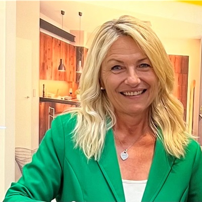 Blonde Frau im grünen Blazer lächelt in moderner, wohnlicher Küchenumgebung.