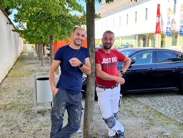 Zwei Handwerker lehnen an einem Baum vor einem Auto auf einem Parkplatz in der Stadt.